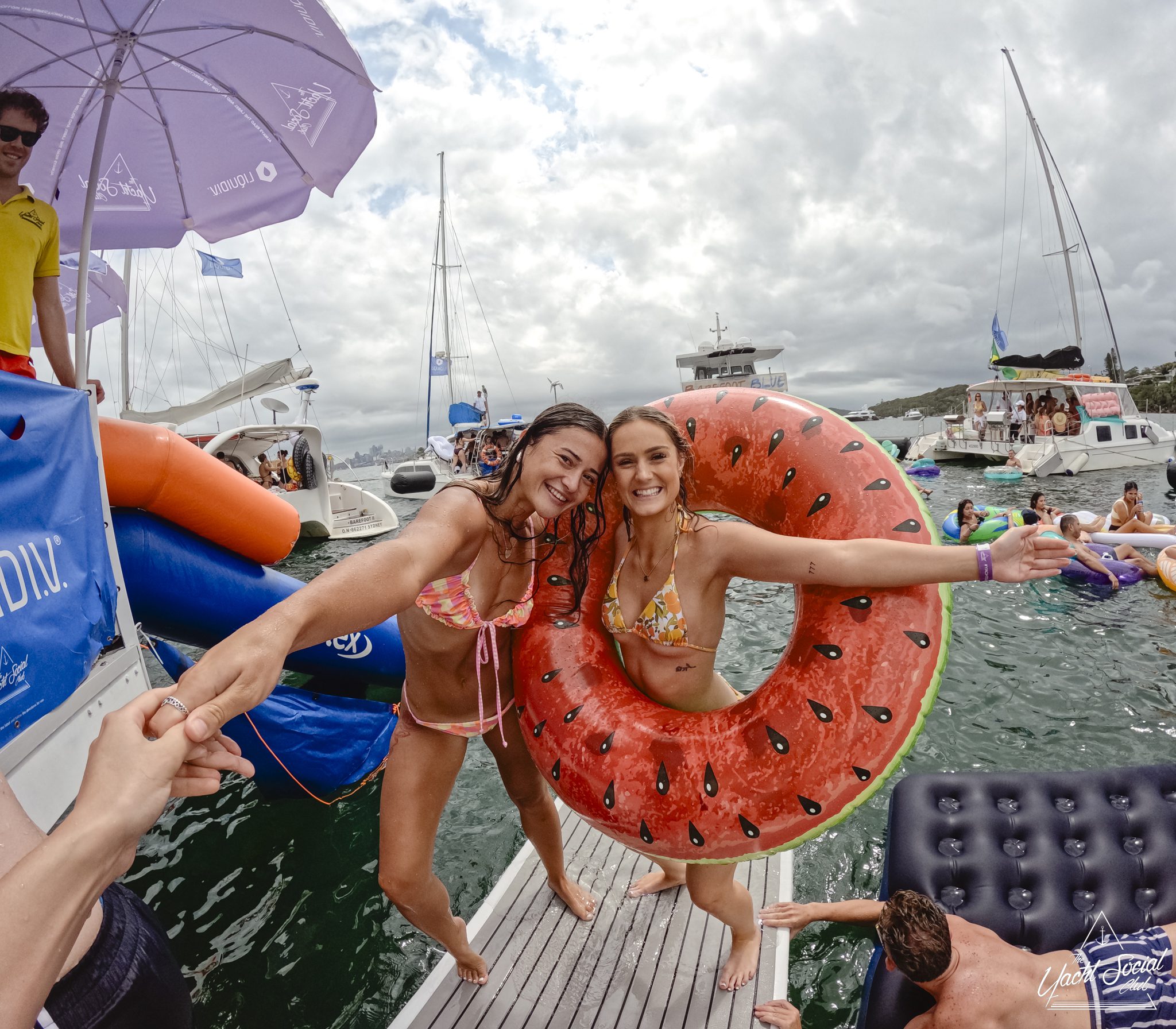 Two smiling women in swimsuits on a boat deck. One has a watermelon-shaped float. They're holding hands with someone off-camera. People and yachts are in the water around them, and a cloudy sky is overhead.
