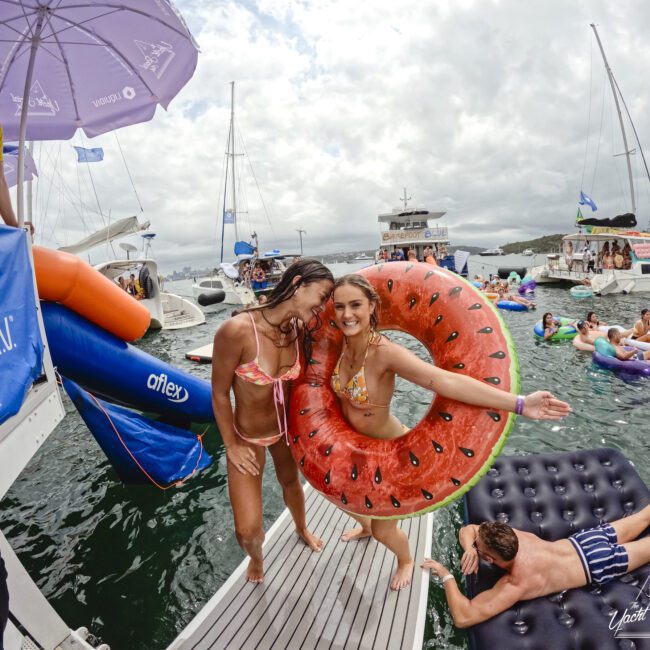 Two women in swimsuits stand on a boat, one wearing a watermelon-shaped float. A man relaxes on an inflatable in the water. In the background, people are enjoying the water near boats under a cloudy sky.