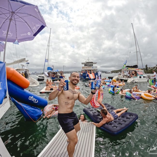A man in swim trunks energetically holds two drink cans on a dock. In the background, people enjoy a festive atmosphere on inflatable floats in the water near boats under a cloudy sky. An umbrella with logos is visible.