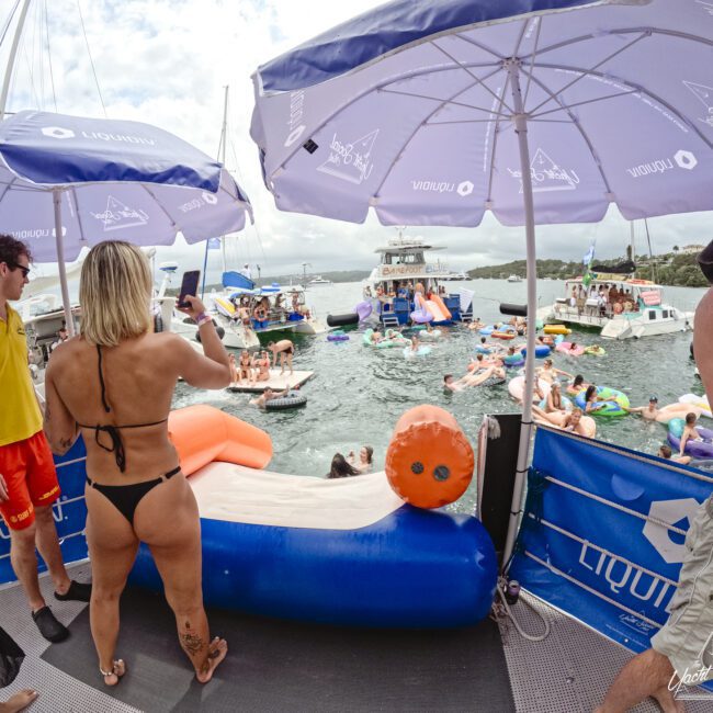 A crowded aquatic party scene featuring people on various inflatable floats in a body of water. A woman in a bikini takes a photo while others relax under umbrellas with a boat visible in the background. Sunny weather adds to the festive atmosphere.