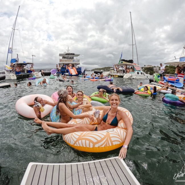 A group of people sitting on colorful inflatable tubes enjoying a party on the water. Boats are anchored nearby, and the sky is cloudy. One person holds a drink, while others smile and relax in the water.