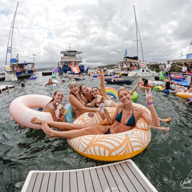 A group of smiling people are lounging on inflatable floaties in the water near boats. One person is holding a drink and making a peace sign. The sky is cloudy, and the scene is festive, with others enjoying the water and boats in the background.
