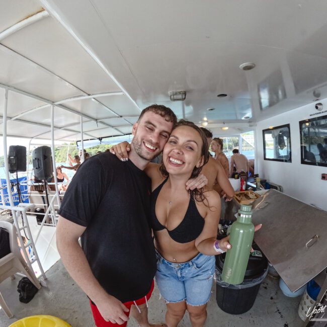A smiling man and woman in casual attire pose for a photo on a boat. The woman is holding a green bottle. Other people are visible in the background, enjoying the boat setting.