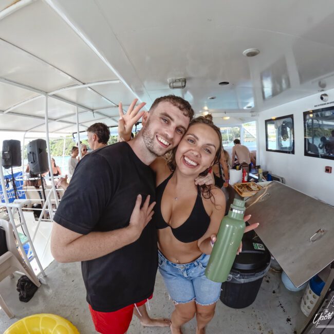 A smiling man and woman pose together on a boat, with the woman holding a green bottle. Other people are gathered in the background. The setting is casual, with chairs and open windows offering a view of the water.