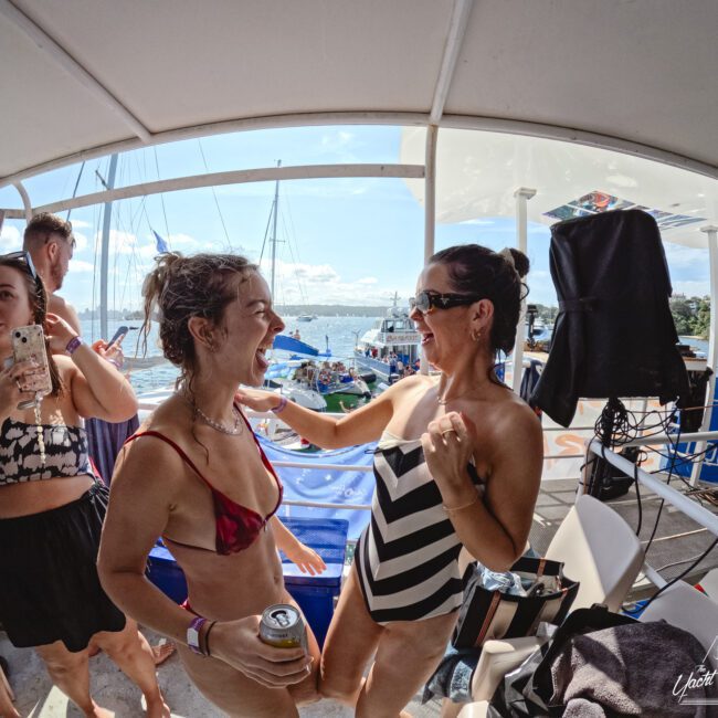 A group of people on a boat are enjoying a sunny day. Two women in swimsuits are laughing and smiling at each other. Other people are holding drinks and taking photos. Sailboats are visible in the background on the water.