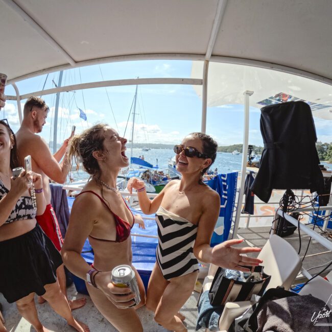 People are enjoying a lively moment on a boat under a canopy. Two women in swimsuits are laughing and dancing with drinks in hand. Others nearby are smiling. Boats and water can be seen in the background on a sunny day.