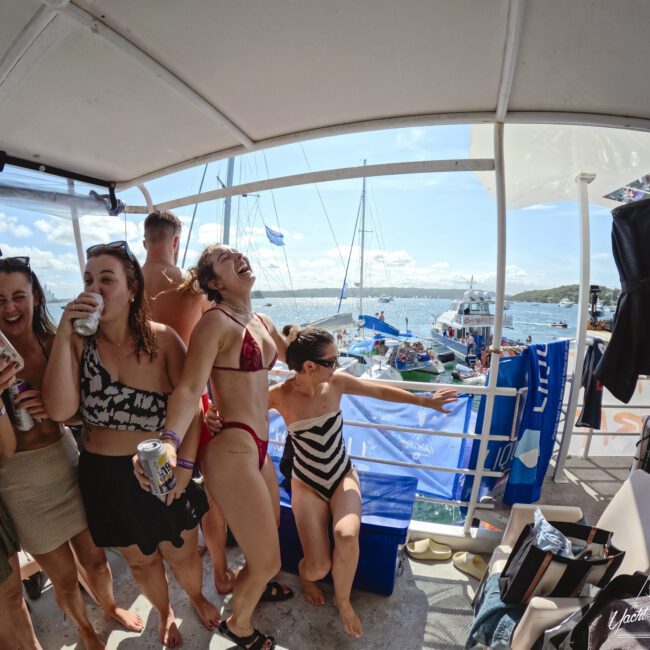 A group of people on a boat enjoying a sunny day. They are laughing, holding drinks, and looking at a phone. The background shows water, another boat, and a blue sky with clouds.