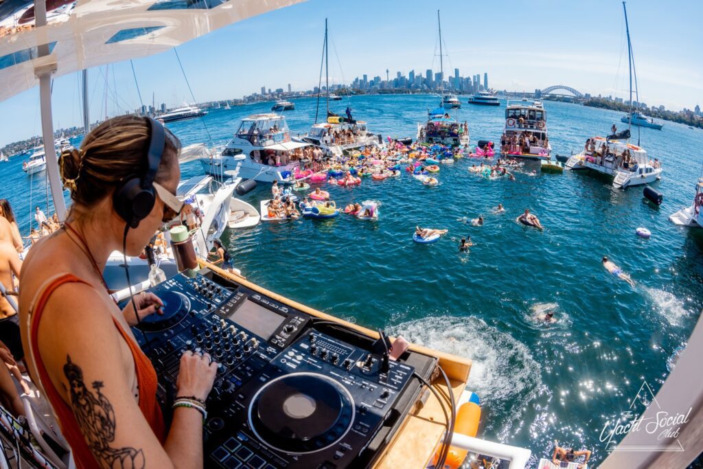 A DJ wearing headphones plays music on a boat overlooking a lively floating party, with people relaxing on inflatable floats and several boats anchored on a sunny harbor, city skyline in the background.