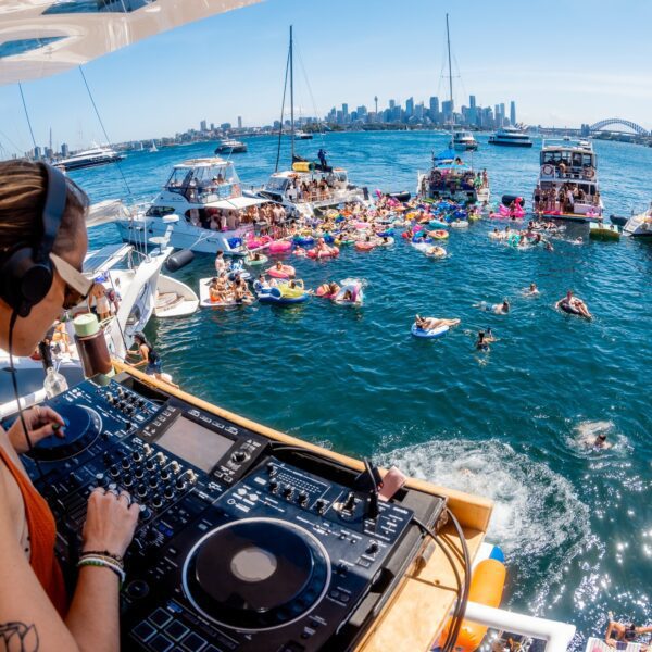 A DJ wearing headphones plays music on a boat overlooking a lively floating party, with people relaxing on inflatable floats and several boats anchored on a sunny harbor, city skyline in the background.