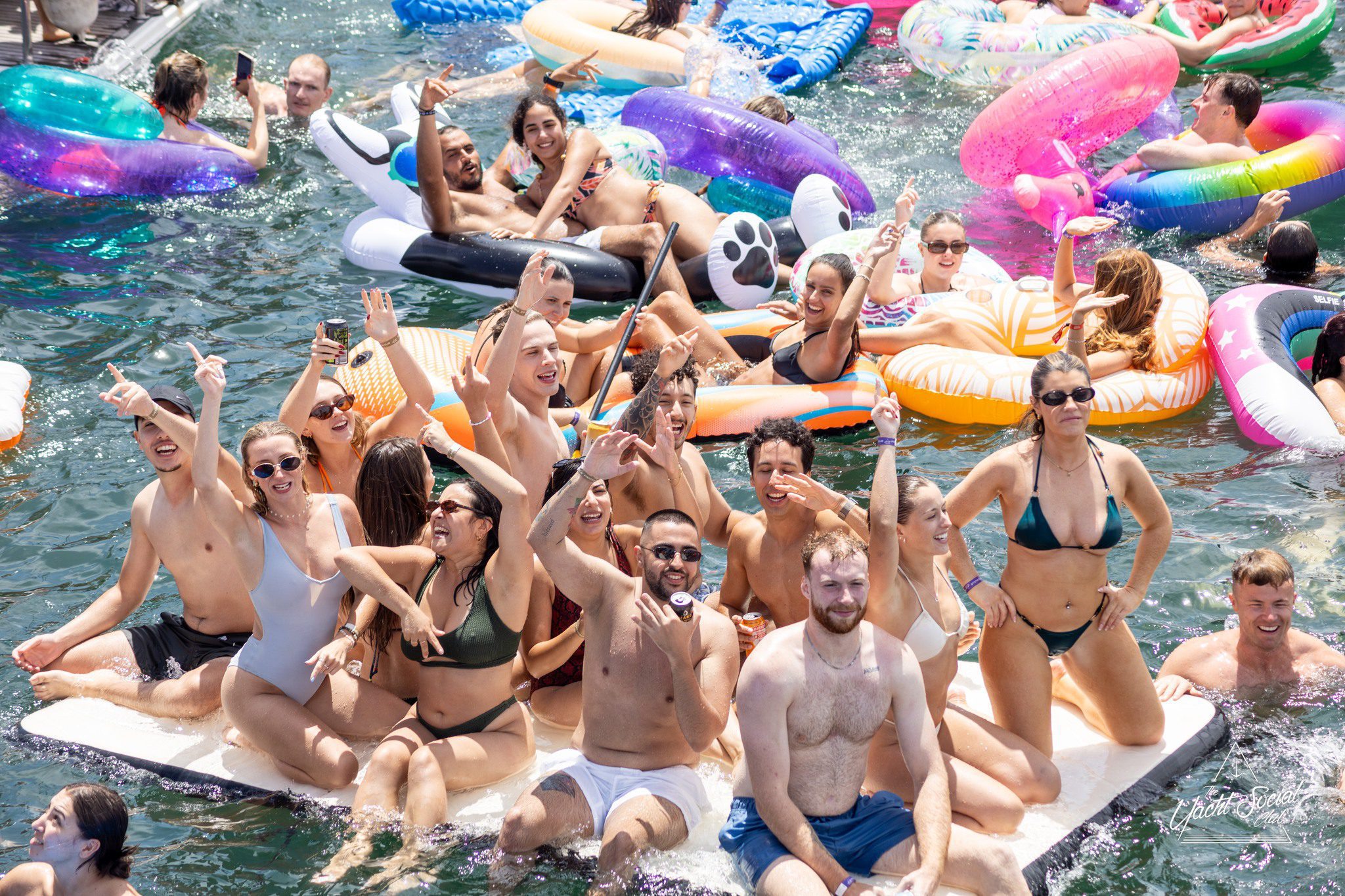 A large group of people in swimsuits smile, cheer, and pose on pool floats and inflatables in the water during a lively summer party. Brightly colored floaties add to the festive atmosphere.