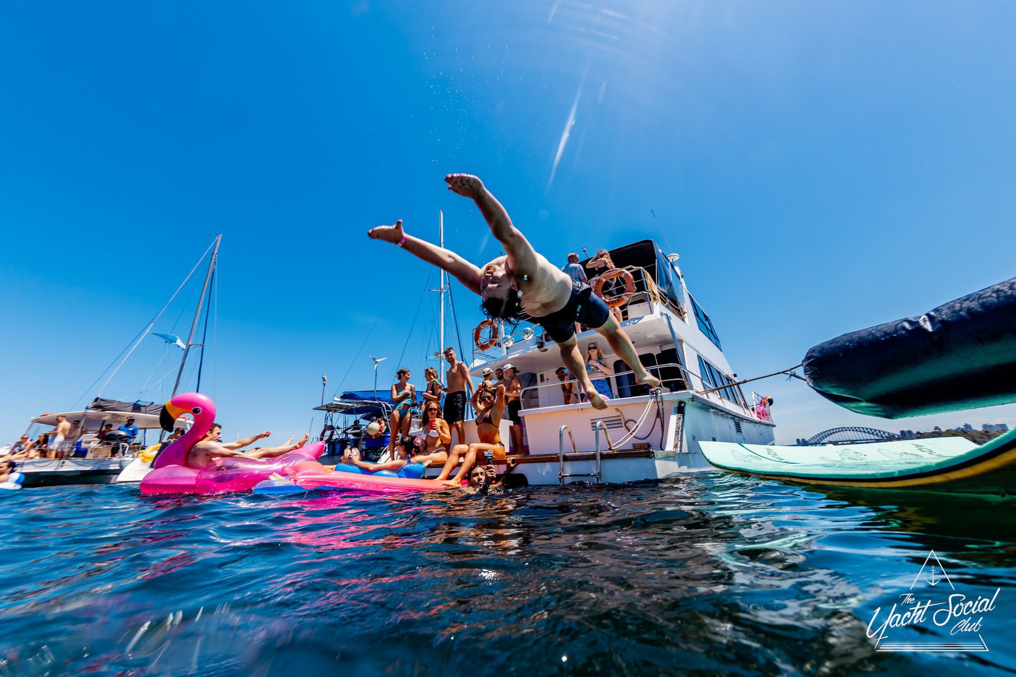 A group of people enjoy a sunny day on yachts. One person dives into the water while others relax, including someone on a large pink flamingo float. The water is calm and the sky is clear and blue.