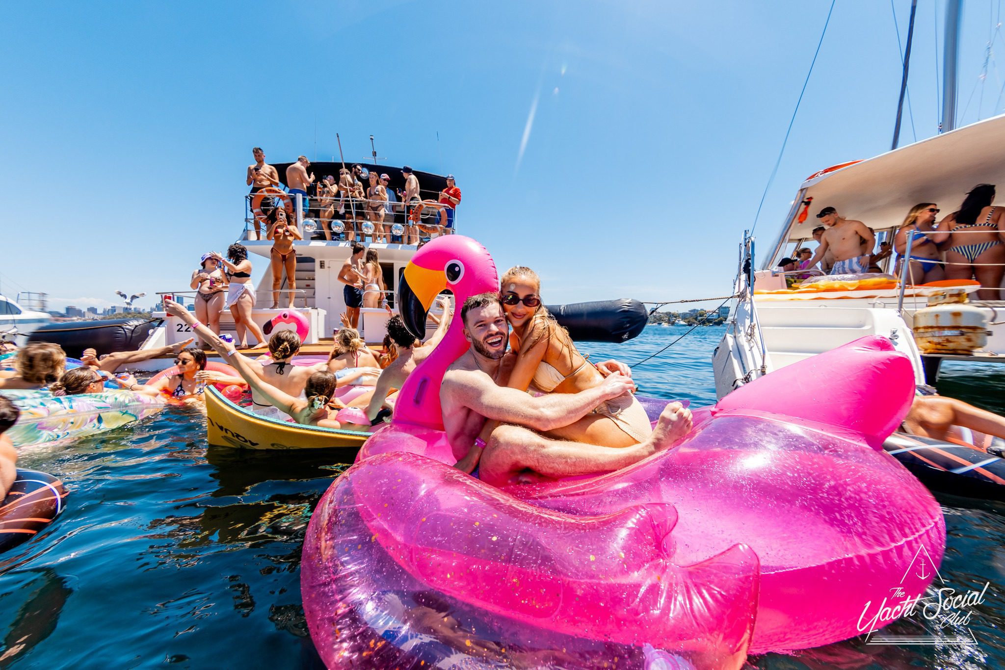 A man and woman smile while sitting on a large pink flamingo float in the water, surrounded by people on other floats and boats, enjoying a sunny group party.