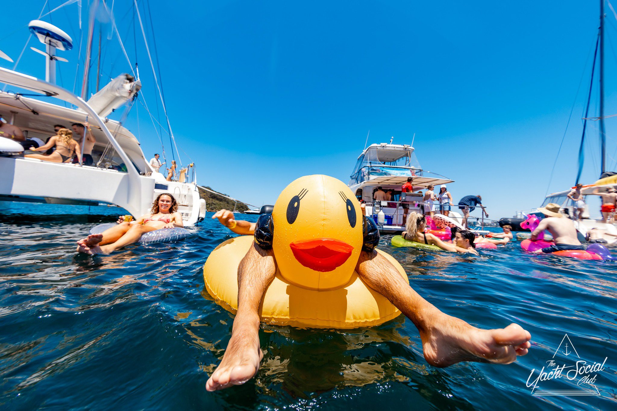 People relax on colorful floats and yachts in a sunny, blue-water setting. A person is featured front and center on a large inflatable duck, their legs stretched out and arms resting on the float.