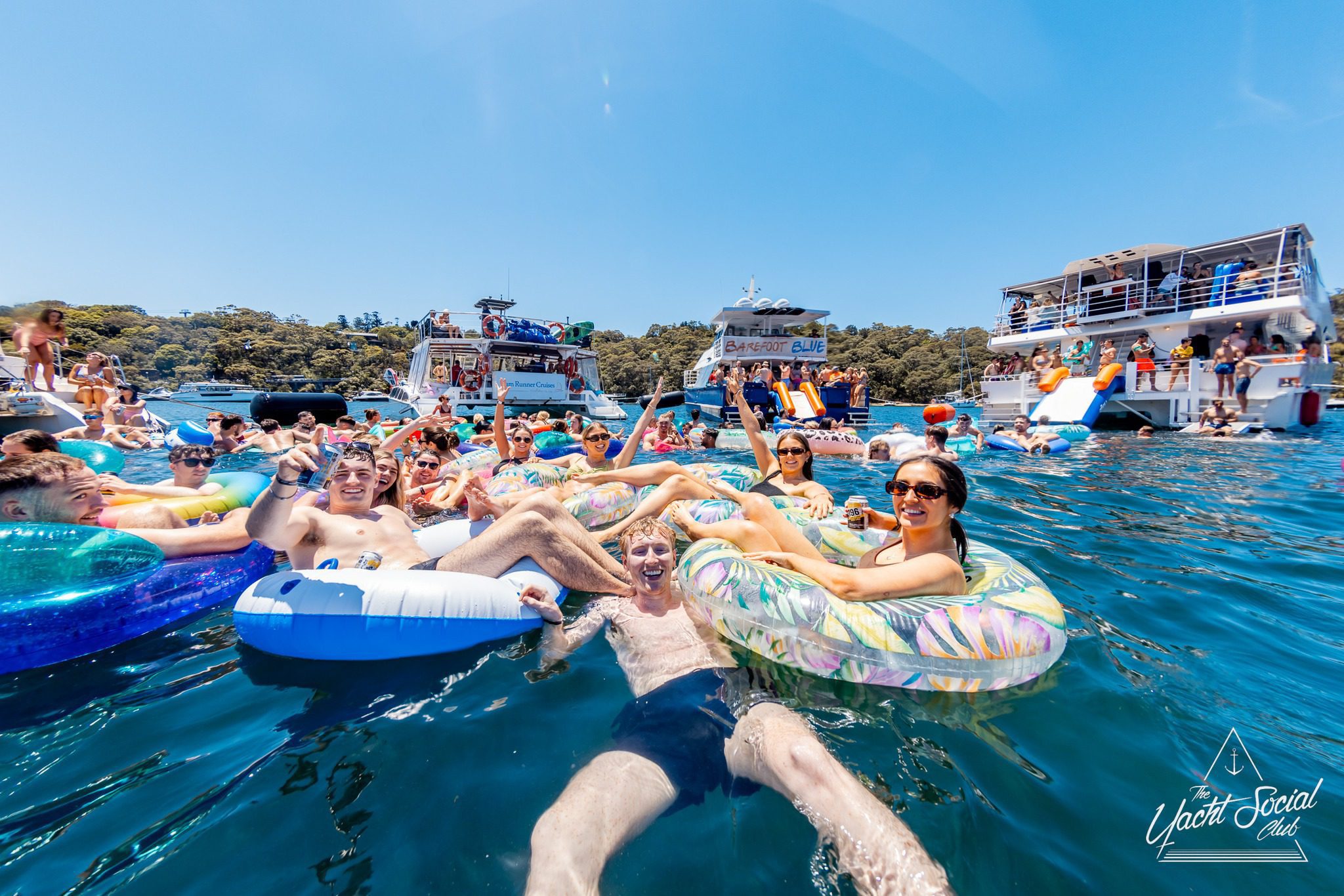 A large group of people relax on colorful pool floats in the water near several yachts, enjoying a lively Yacht Club gathering. The scene is festive, with blue skies and green trees in the background.