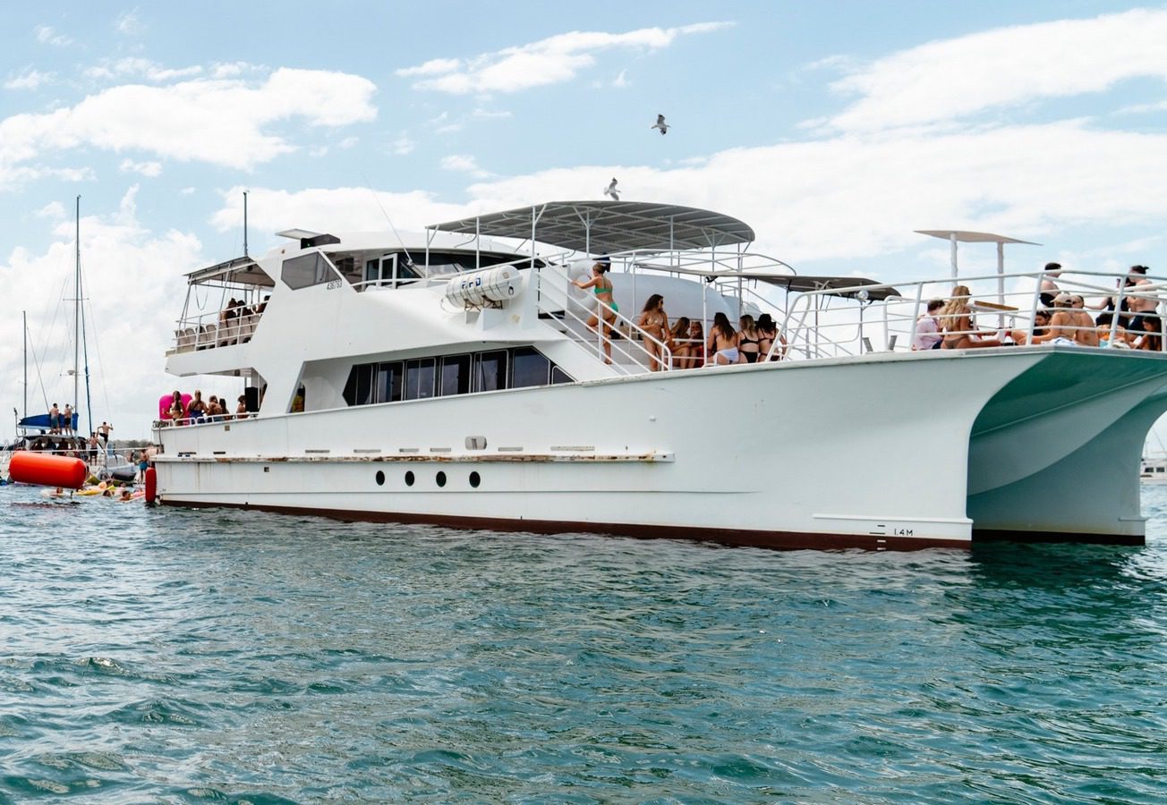 A large white catamaran yacht with people on board is anchored on blue water under a partly cloudy sky; smaller boats and people swimming are visible nearby.