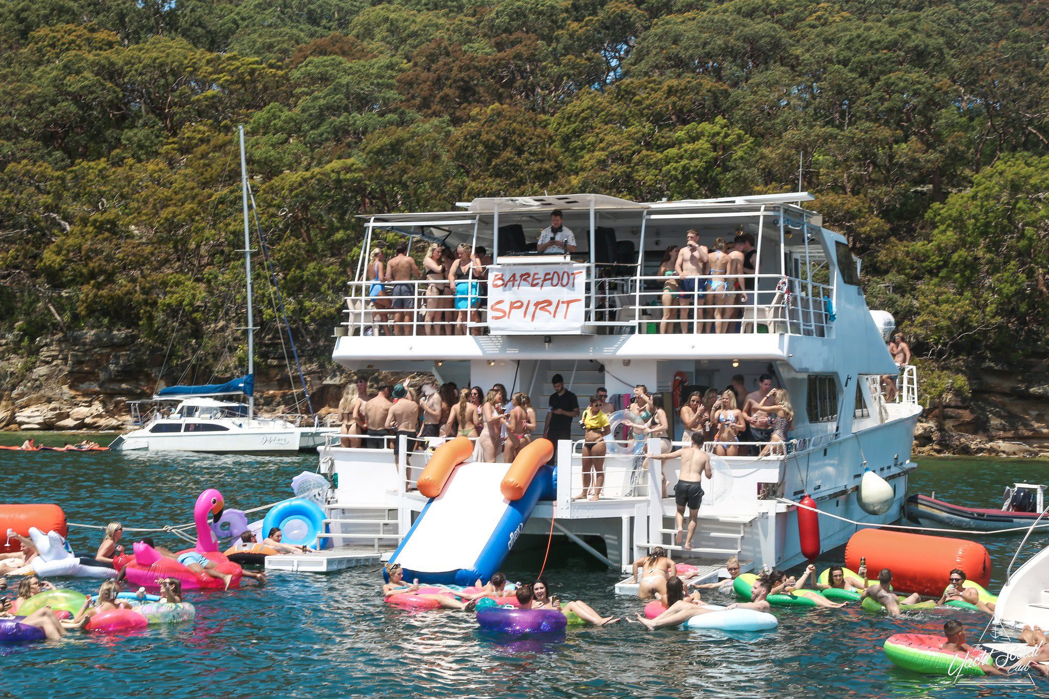 A large group of people enjoy a party on a docked boat called "Barefoot Spirit," with others on colorful inflatables in the water nearby. A forested shoreline and another boat are visible in the background.