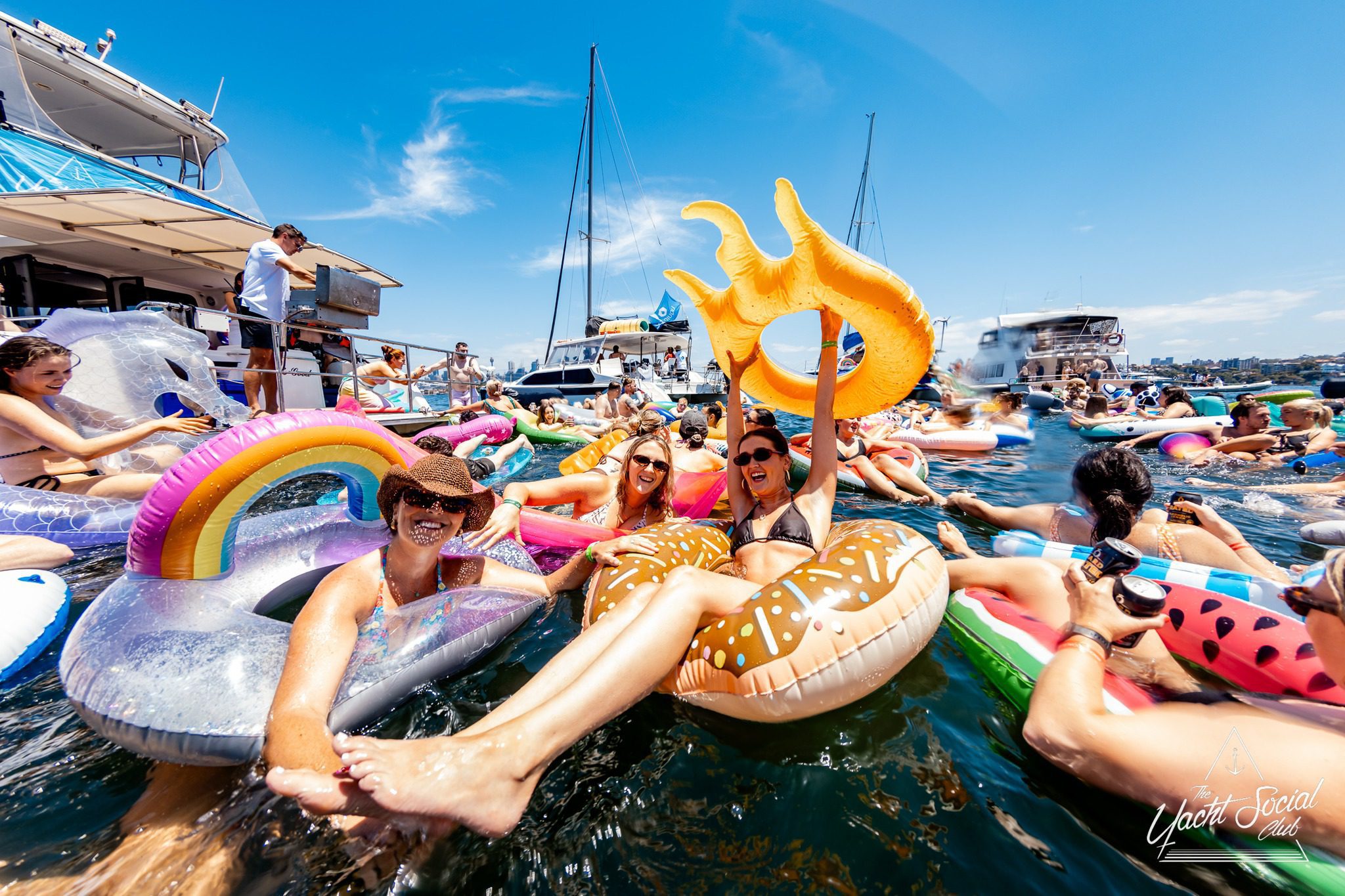 People are relaxing on colorful inflatable pool floats in the water near yachts on a sunny day, smiling and enjoying a festive atmosphere. The scene is lively with blue skies and party energy.