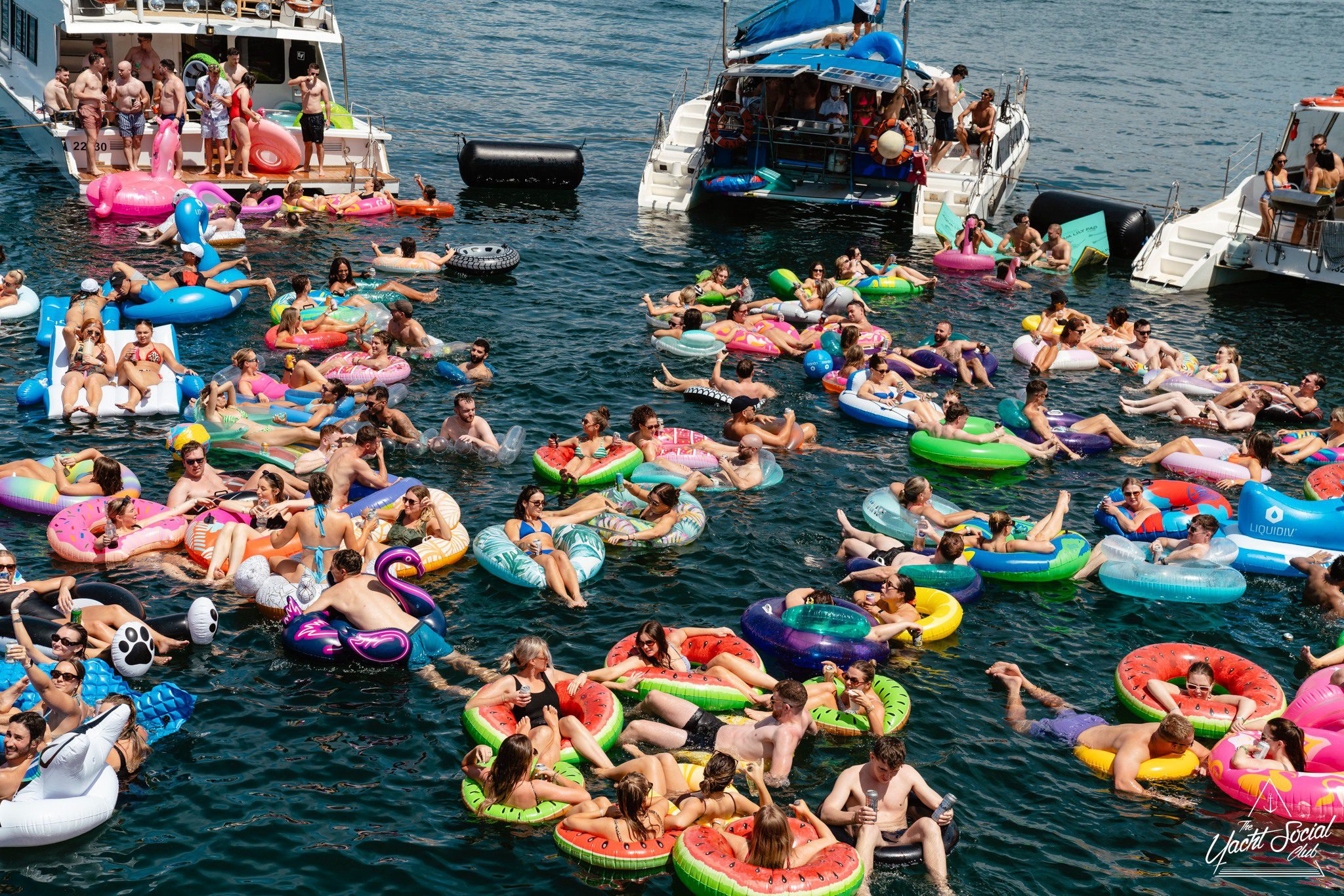 A large group of people relax on colorful inflatable floats in the water near two docked boats on a sunny day, enjoying a lively summer gathering.