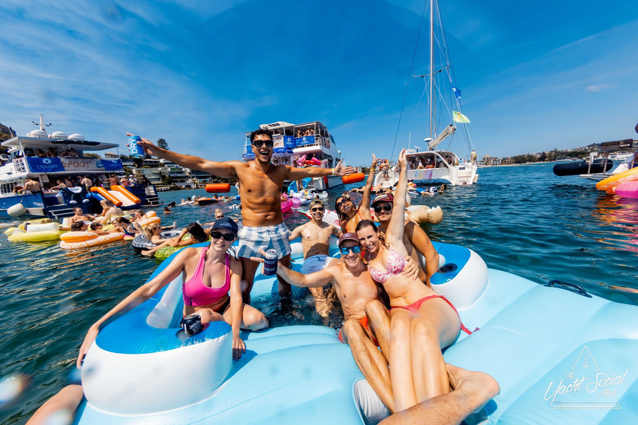A group of young adults in swimsuits relax and smile on a large inflatable float in the water, with boats and other people on floats in the background under a sunny blue sky.