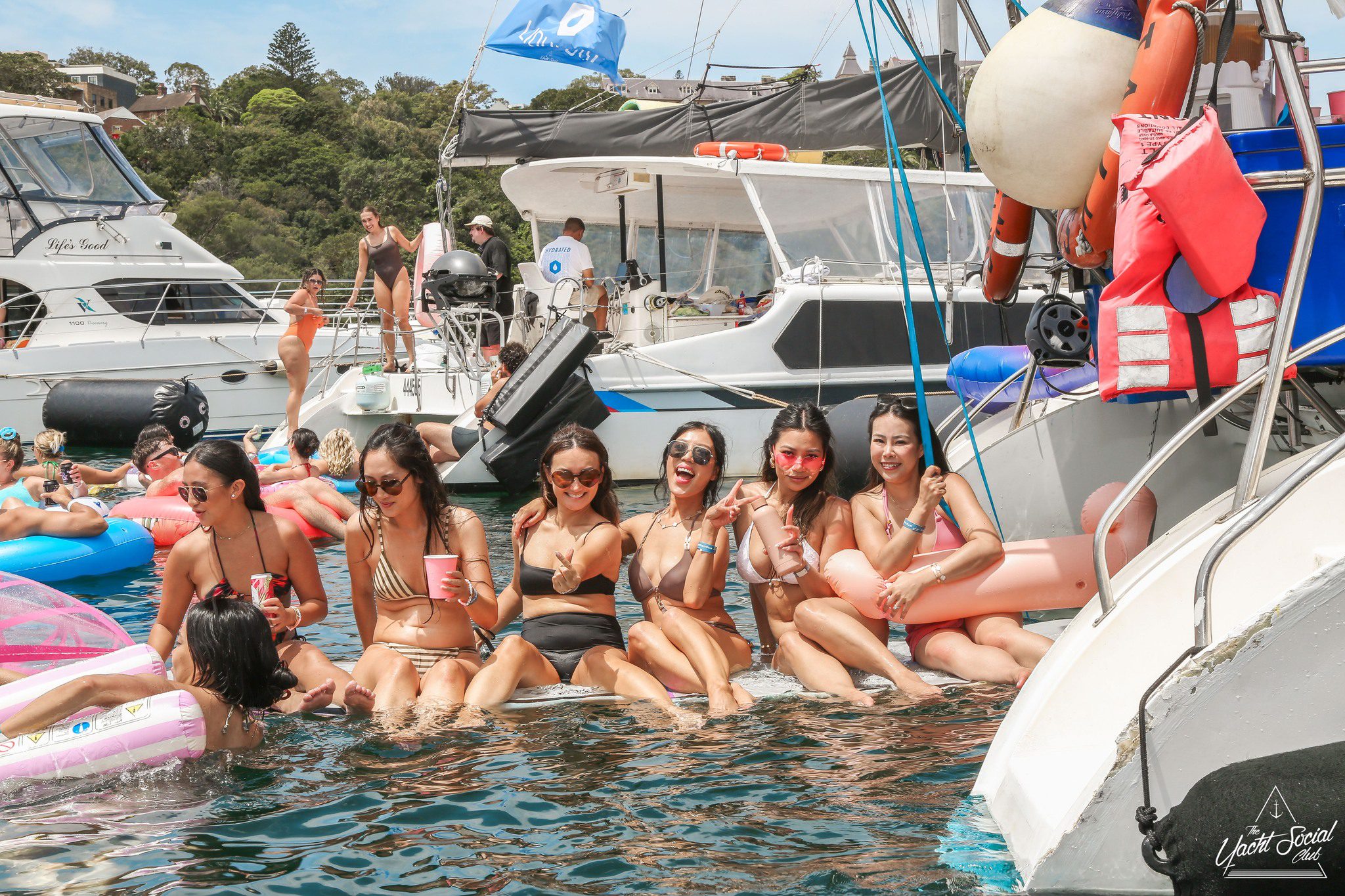 Six women in swimsuits sit laughing on the edge of a boat, their feet in the water. Other people and boats surround them. It is a sunny day, and the atmosphere appears festive and relaxed.