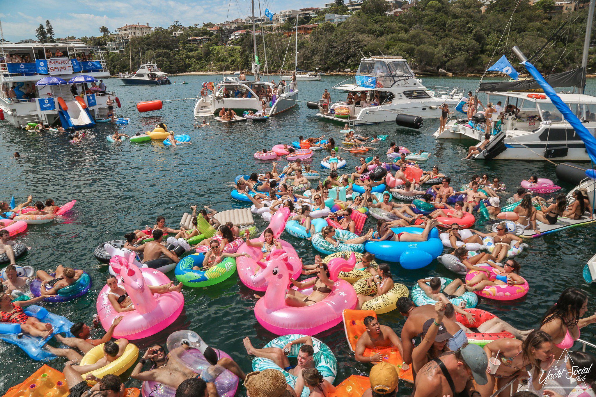 A large crowd of people relax on colorful inflatable pool floats in the water near several anchored boats on a sunny day, surrounded by green hills and waterfront homes.