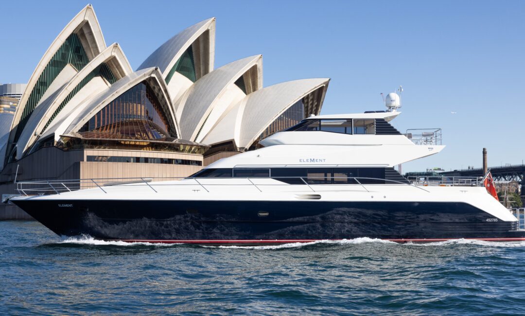 A luxury yacht sails on blue water in front of the Sydney Opera House under a clear sky.