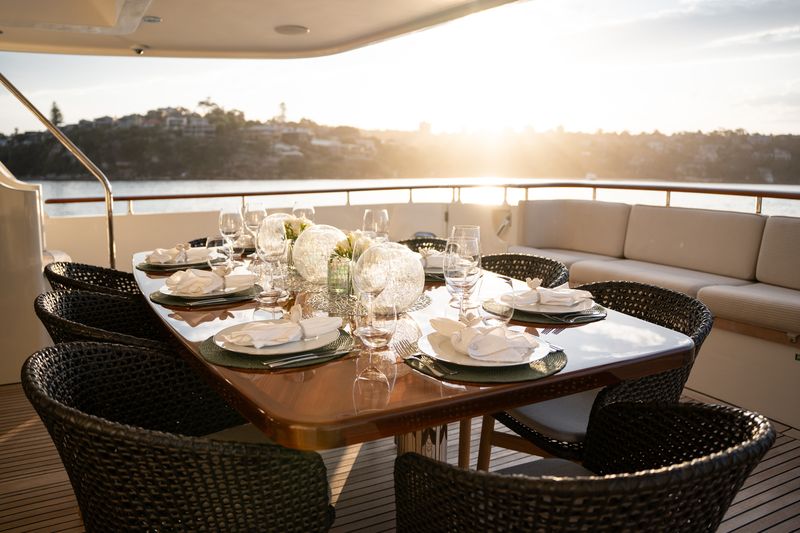 Elegant outdoor dining table set for six on a yacht, featuring white flower centerpieces, glassware, and plates, with sunlight reflecting off the water and a scenic shoreline in the background.