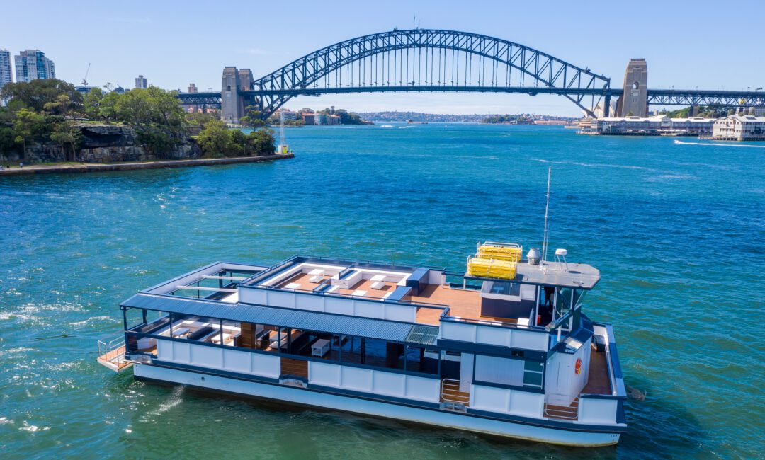 A modern houseboat floats on blue water with the Sydney Harbour Bridge in the background under a clear sky, and city buildings and greenery visible along the shoreline.
