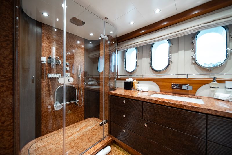 Luxurious yacht bathroom with marble surfaces, a glass-enclosed shower, wood cabinetry, large countertop, and three round porthole windows letting in natural light. Towels and toiletries are neatly arranged.