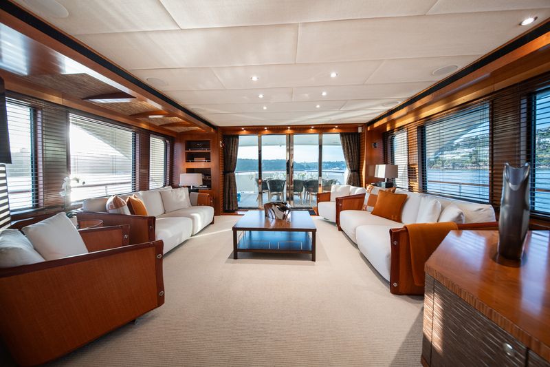Luxurious yacht interior with large windows, wooden paneling, white sofas, a central coffee table, and outdoor seating area visible through glass doors overlooking the water.