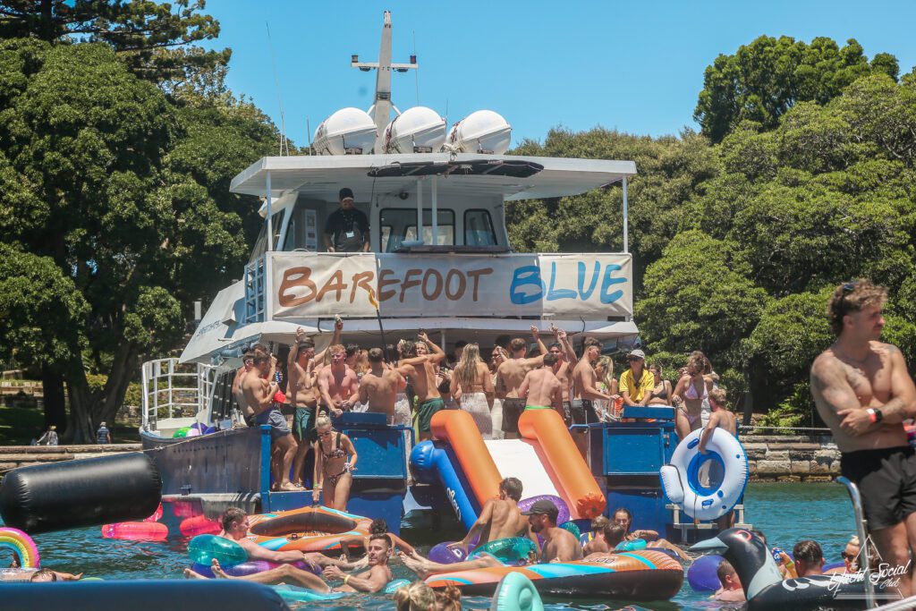A large boat with a "Barefoot Blue" banner hosts a lively party, with many people in swimsuits dancing and cheering. Colorful floaties and inflatable slides are in the water; trees line the background.