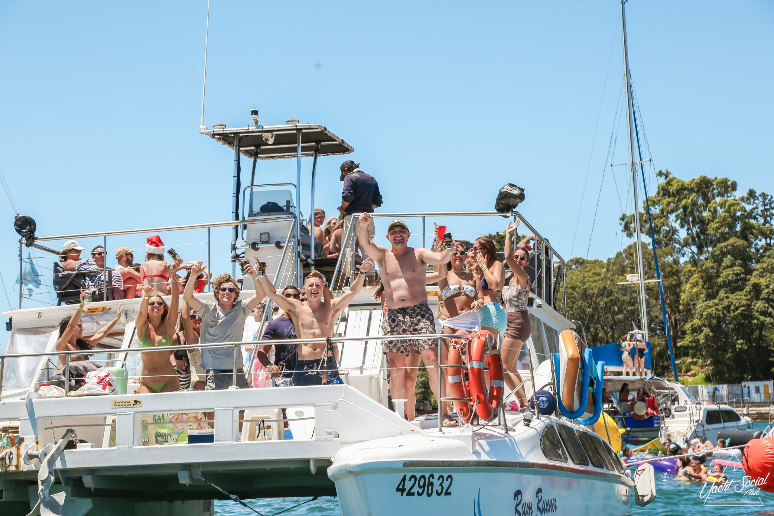A group of people in swimwear cheer and raise their arms while standing on a boat. The scene is sunny, festive, and the boat is surrounded by water with other boats and people in the background.
