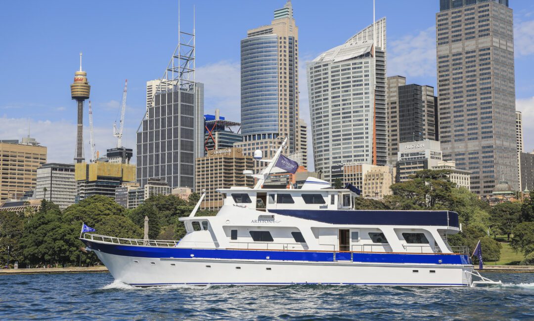 A white yacht with a blue stripe sails on the water, with a city skyline of modern high-rise buildings and a communications tower in the background, under a clear blue sky.