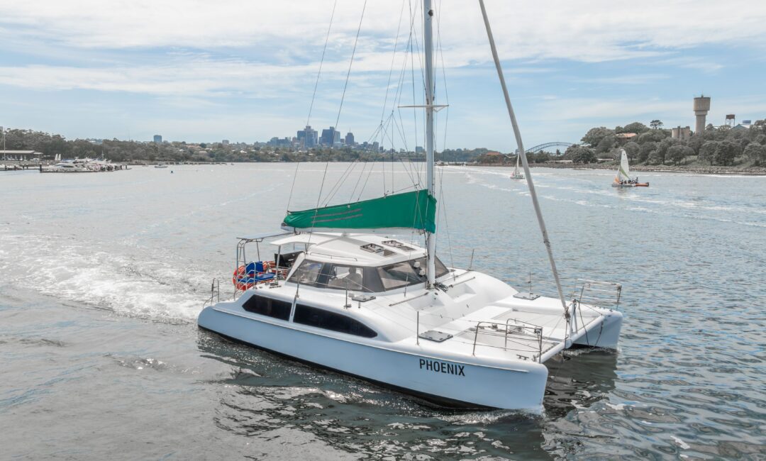 A white catamaran named "PHOENIX" sails on a calm body of water with a green canopy and city skyline in the background under a partly cloudy sky.