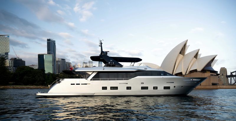 A sleek, modern yacht floats on calm water near the iconic Sydney Opera House, with city buildings and a partly cloudy sky in the background.