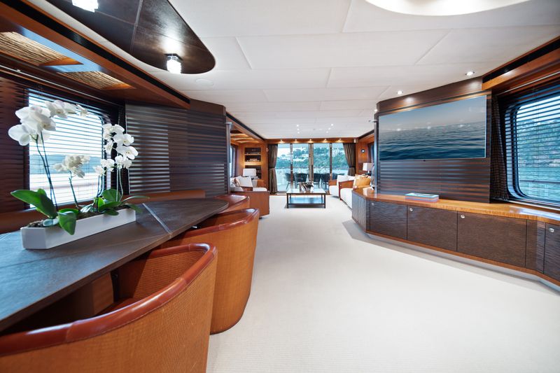 Modern yacht interior with large windows, wooden panels, and tan leather chairs. A white orchid sits on a counter. There is a wall-mounted TV and a spacious seating area with natural light filling the room.