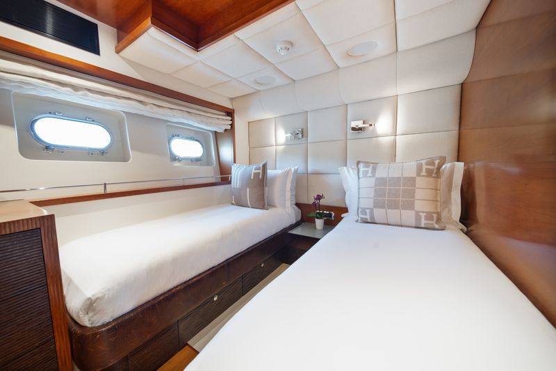 A cozy yacht cabin with two single beds, each with white bedding and decorative pillows, wood accents, two small windows, and soft, padded wall panels. A small table with a plant is placed between the beds.