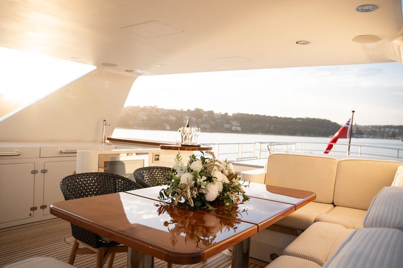 A polished wooden table with a floral arrangement sits on the deck of a yacht, surrounded by cream cushions and wicker chairs, with a view of water and shoreline in the background.