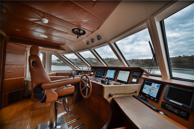 The interior of a modern yacht's helm featuring a brown captain’s chair, multiple navigation screens, control panels, and large windows offering a view of water and a distant shoreline.