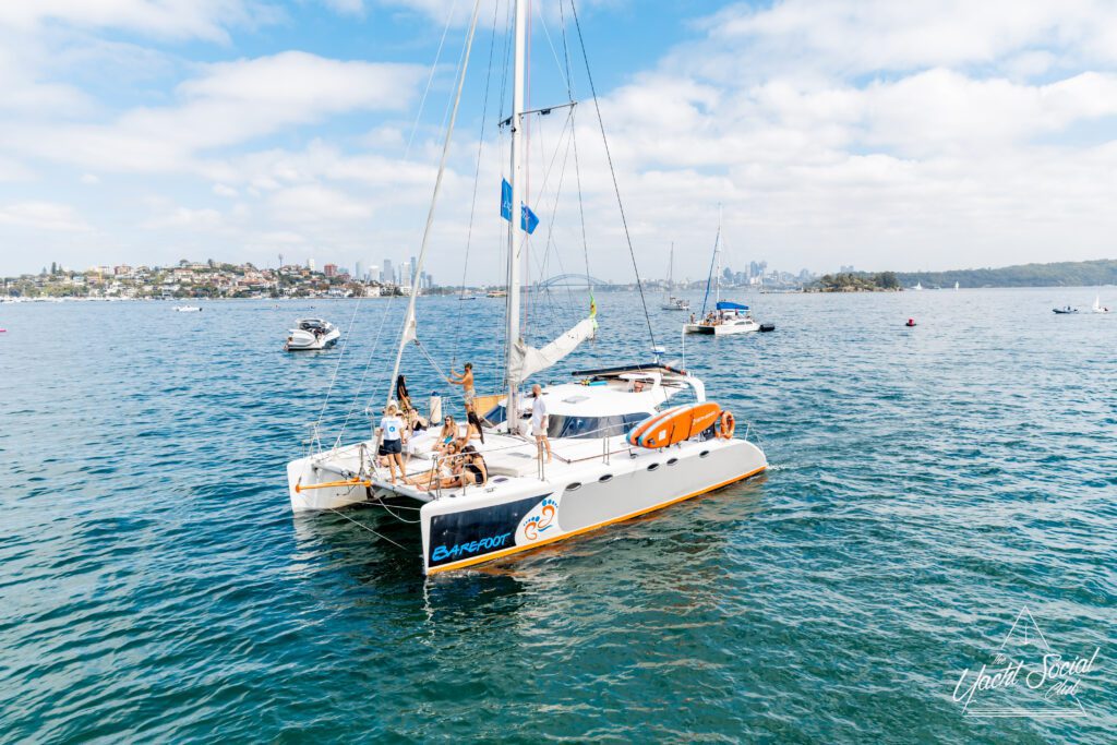 A white catamaran with several people on board sails on calm blue water near a city skyline, with other boats nearby under a partly cloudy sky.