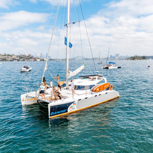 A white catamaran with several people on board sails on calm blue water near a city skyline, with other boats nearby under a partly cloudy sky.