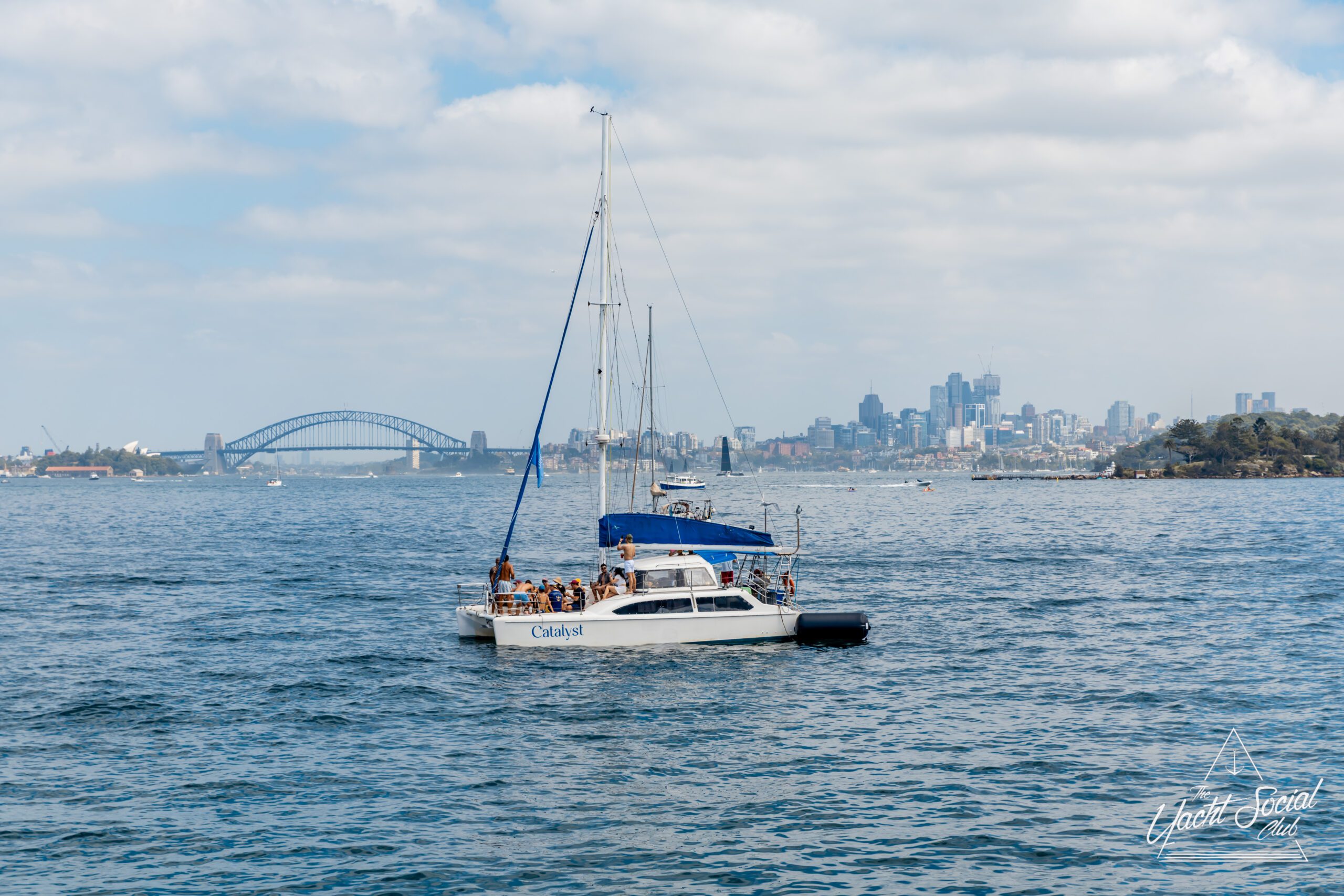 A white catamaran with people onboard sails on Sydney Harbour, with the Sydney Harbour Bridge and city skyline visible in the background under a partly cloudy sky.