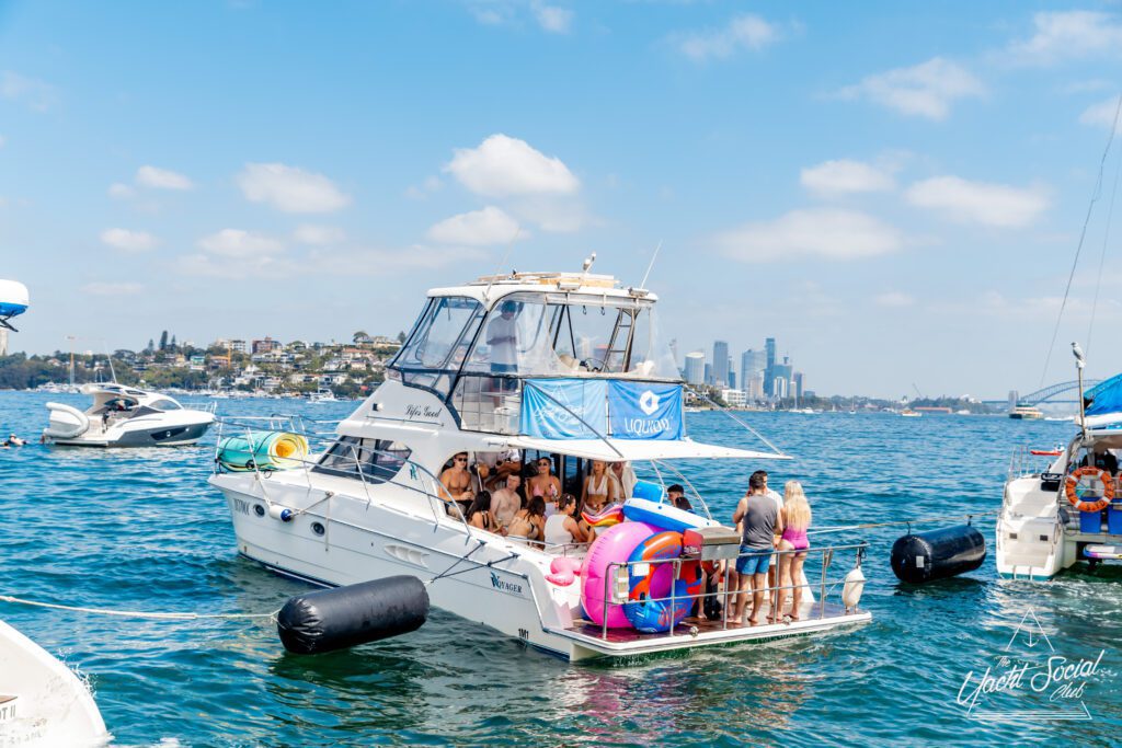 A group of people in swimwear gather on the deck of a white yacht decorated with a blue flag, floating on a sunny day with city buildings visible in the distant background. Other boats are nearby on the water.