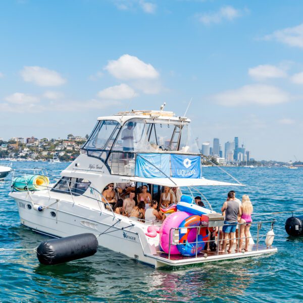 A group of people in swimwear gather on the deck of a white yacht decorated with a blue flag, floating on a sunny day with city buildings visible in the distant background. Other boats are nearby on the water.
