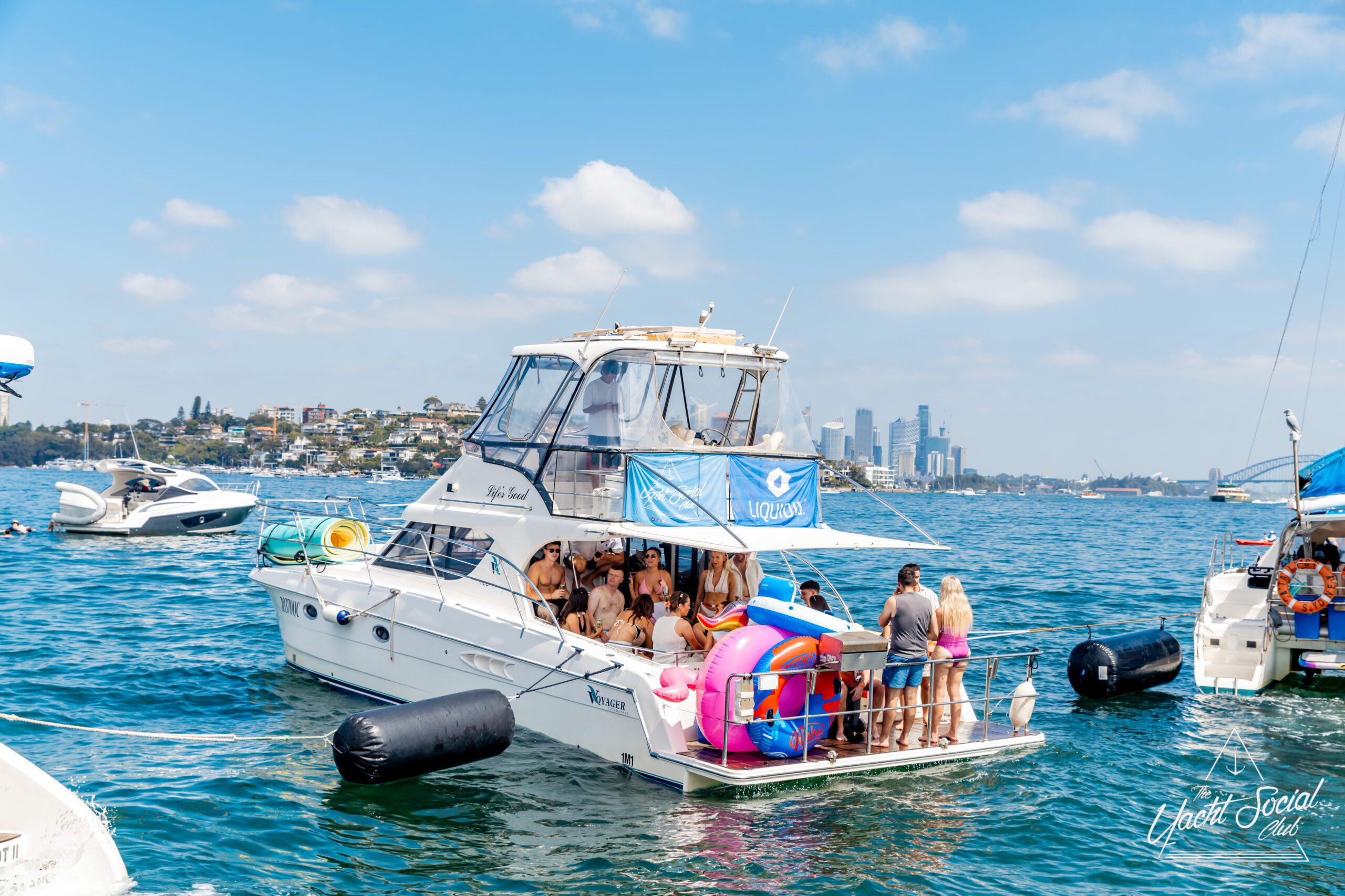 A group of people in swimwear gather on the deck of a white yacht decorated with a blue flag, floating on a sunny day with city buildings visible in the distant background. Other boats are nearby on the water.