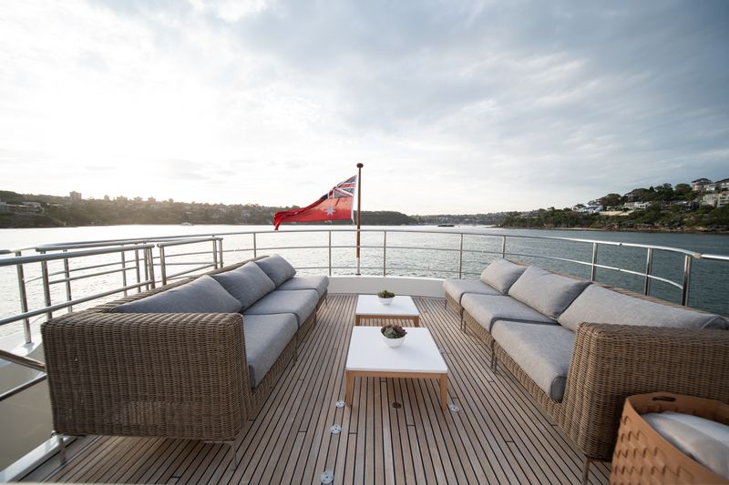 Two wicker sofas with gray cushions face each other on a yacht deck, with a white table between them. A red maritime flag waves at the stern, and water and shoreline are visible in the background.