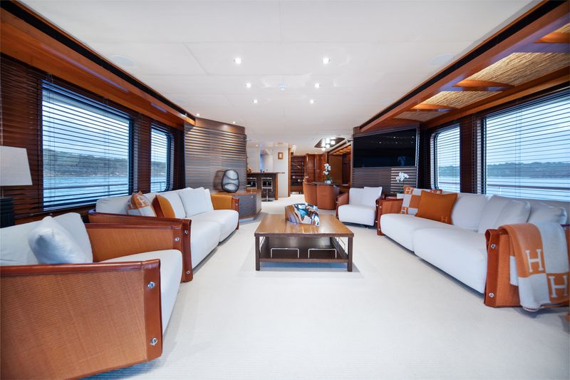Spacious, modern yacht interior with white sofas, wooden accents, large windows with blinds, and a central coffee table. Bright, natural light fills the room, offering views of the water outside.