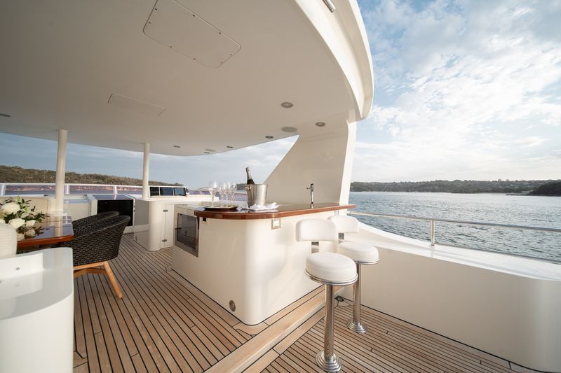 Luxury yacht deck with wooden flooring, a modern bar area with stools, a bottle of champagne in an ice bucket, a table with flowers, and a scenic view of water and distant shoreline under a partly cloudy sky.
