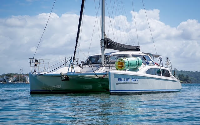 A white catamaran sailboat named "Blue Sky" floats on calm blue water under a partly cloudy sky, with green tanks secured on its deck and a coastal landscape visible in the background.