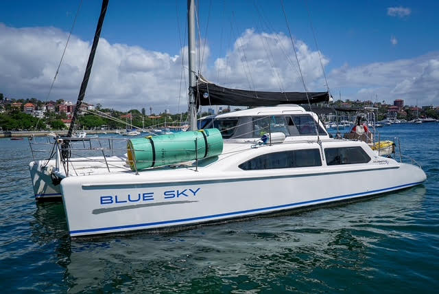 A white catamaran named "BLUE SKY" floats on calm water near a marina under a partly cloudy sky, with green tanks secured on its deck and buildings visible in the background.